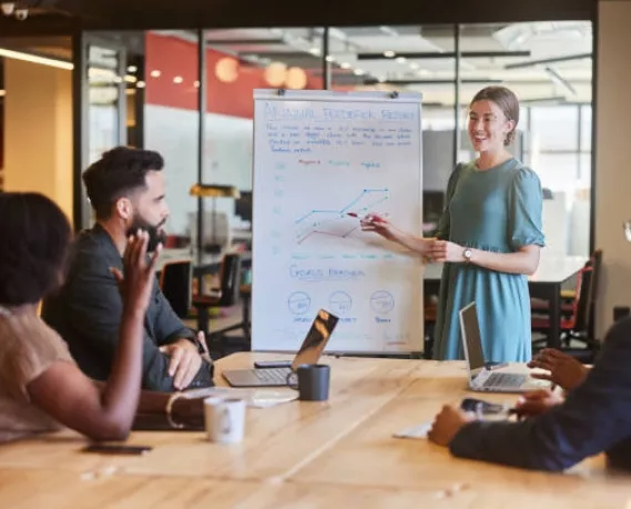 istockphoto-1475863848-612×612 Smiling young businesswoman giving a presentation on a whiteboard to a diverse group of colleagues sitting in an office